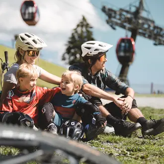 Familie mit Helmen und Mountainbikes sitzt auf einer Wiese vor einer Seilbahn