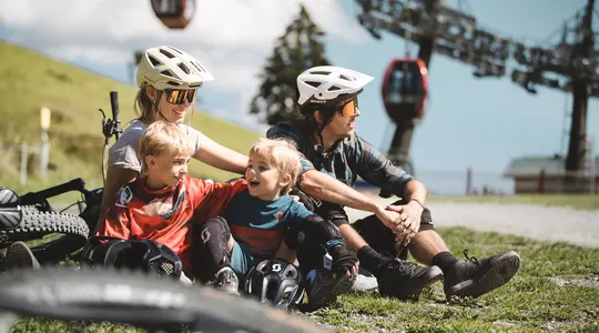 Familie mit Helmen und Mountainbikes sitzt auf einer Wiese vor einer Seilbahn