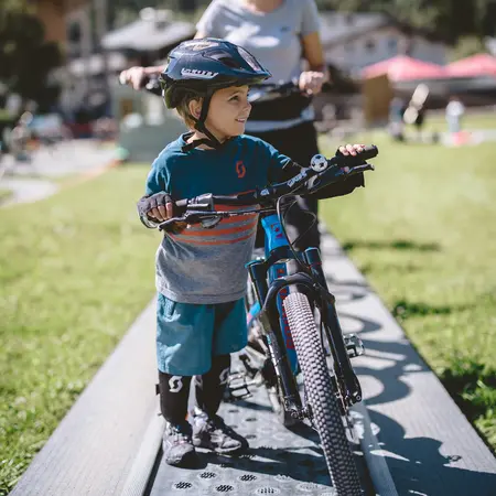 Kind mit Helm fährt Fahrrad auf einem Übungsparcours, Mutter im Hintergrund