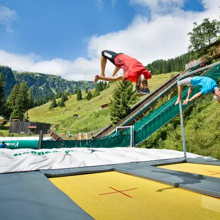 Zwei Jungen springen auf Trampolinen vor Bergkulisse und machen Saltos