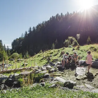 Familie sitzt an Quelle im Grünen vor bewaldetem Berghang unter strahlender Sonne