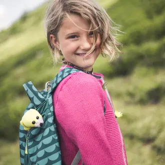 Lächelndes Mädchen mit Rucksack und Kuscheltier vor grünem Hügel