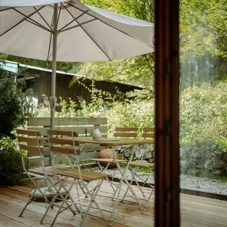 Holzterrasse mit Tisch, Stühlen und Sonnenschirm vor üppigem Grün