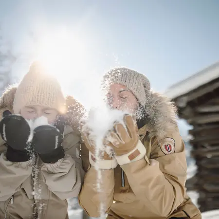 Zwei Personen pusten Schnee in der Wintersonne, warme Kleidung und Mützen