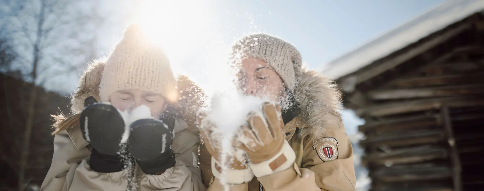Zwei Personen pusten Schnee in der Wintersonne, warme Kleidung und Mützen