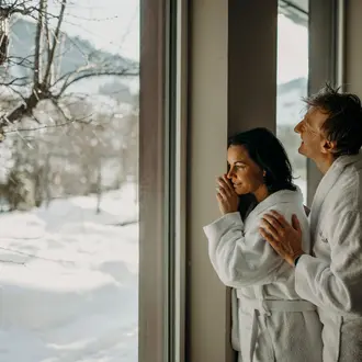 Paar in Bademänteln blickt aus Fenster auf verschneite Winterlandschaft
