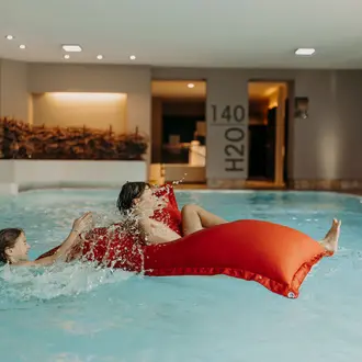 Two people in a swimming pool, one on a red air mattress, splashing water at each other