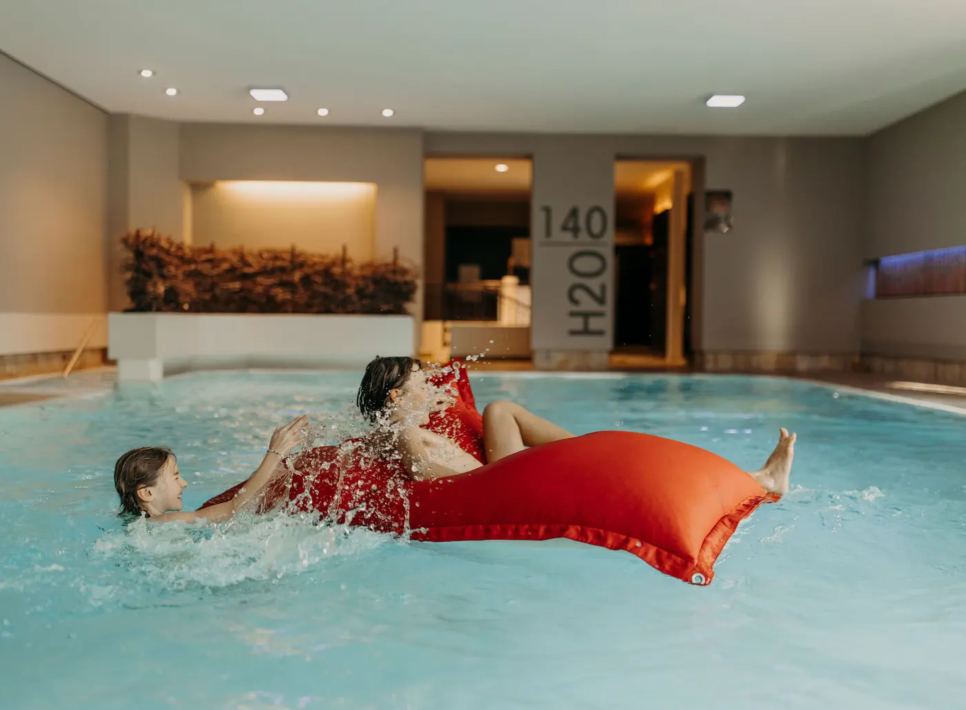 Zwei Personen im Schwimmbad, eine auf roter Luftmatratze, spritzen sich gegenseitig mit Wasser an