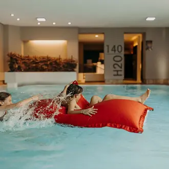 Frau auf rotem Kissen im Schwimmbad, andere Person zieht Kissen ins Wasser