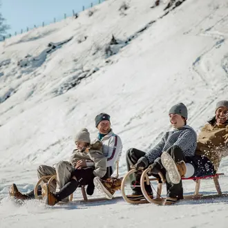Vier Personen fahren auf Holzschlitten einen schneebedeckten Hang hinunter