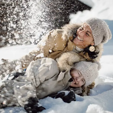 Frau und Kind lachen im Schnee, mit Mützen und Winterjacken bekleidet