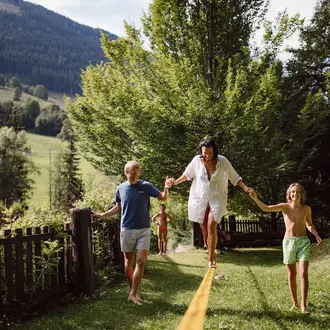 Familie balanciert barfuß auf einem gelben Band im Garten vor Bergkulisse