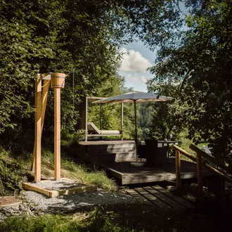 Holzterrasse mit Liege, Tisch und Sonnenschirm im Grünen, Dusche aus Holz im Vordergrund