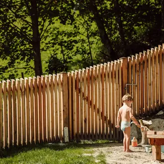 Kind steht vor Holzzaun im Garten und spielt mit Wasserhahn