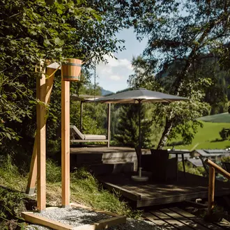 Holzschauerdusche im Freien mit Eimer, Liege und Sonnenschirm auf Holzterrasse vor grüner Landschaft