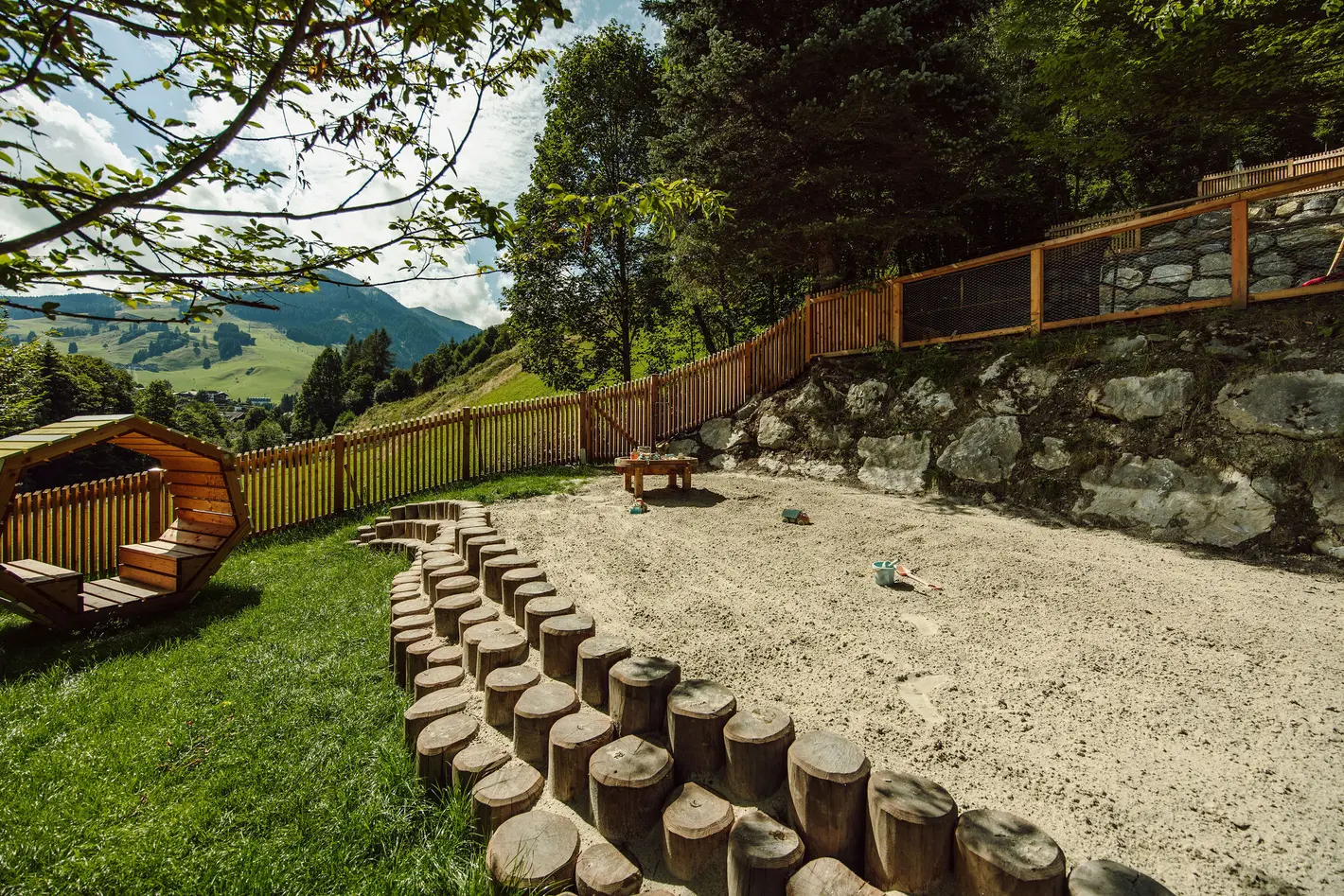 Sandkasten mit Baumstamm-Begrenzung, Holzzaun, Tisch und Bergen im Hintergrund