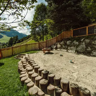 Sandkasten mit Baumstamm-Begrenzung, Holzzaun, Tisch und Bergen im Hintergrund