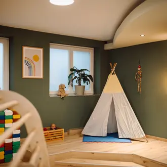 Kinderzimmer mit Tipi, Fenster, Pflanze, Spielzeug und Leiter vor grüner Wand