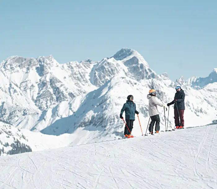 Drei Skifahrer auf schneebedecktem Hang vor Bergpanorama unter blauem Himmel