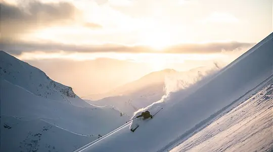 Skifahrer fährt steilen Hang hinab, Staubwolke, Bergpanorama im goldenen Abendlicht